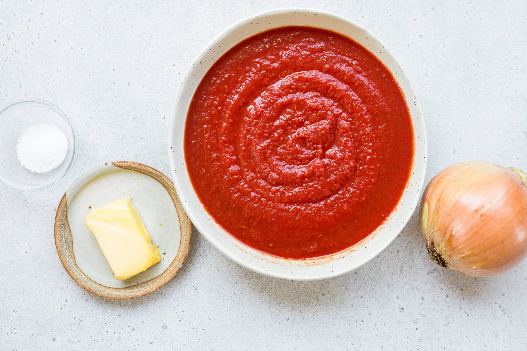 Ingredients for tomato sauce including canned tomatoes, butter, and onion on a white surface
