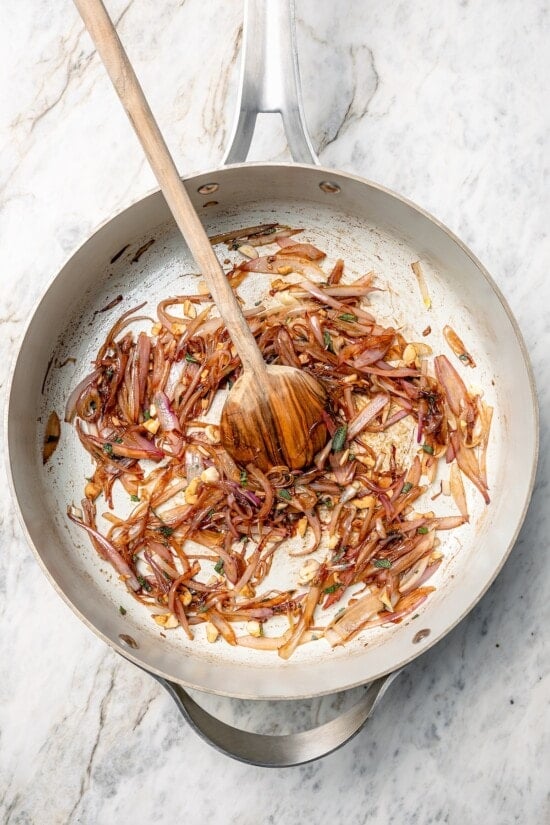 Baked butternut squash is layered with creamy goat cheese, shallots, and pistachios in this cozy side dish recipe. Perfect for the holidays! Shallots cooking in skillet with wooden spoon.