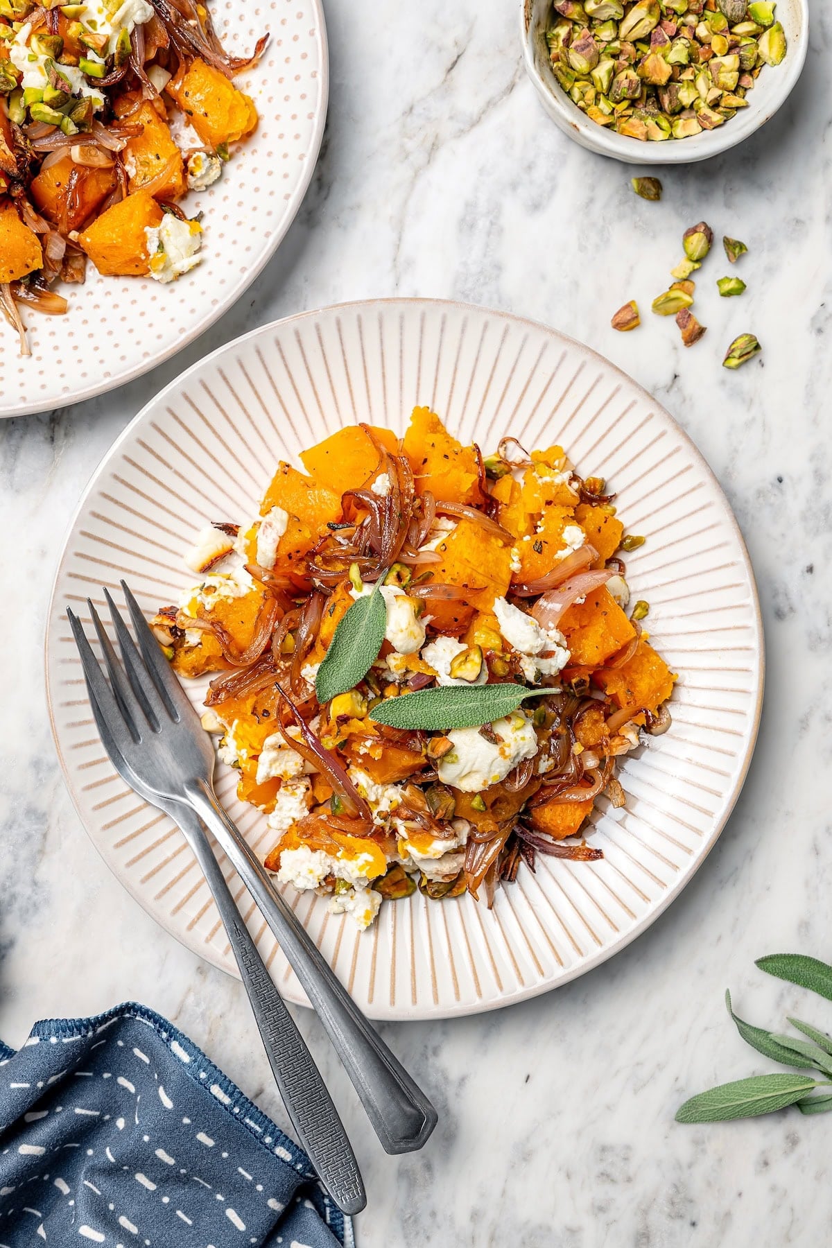 Baked butternut squash is layered with creamy goat cheese, shallots, and pistachios in this cozy side dish recipe. Perfect for the holidays! Baked butternut squash on 2 plates, topped with goat cheese, pistachios, shallots, and sage.