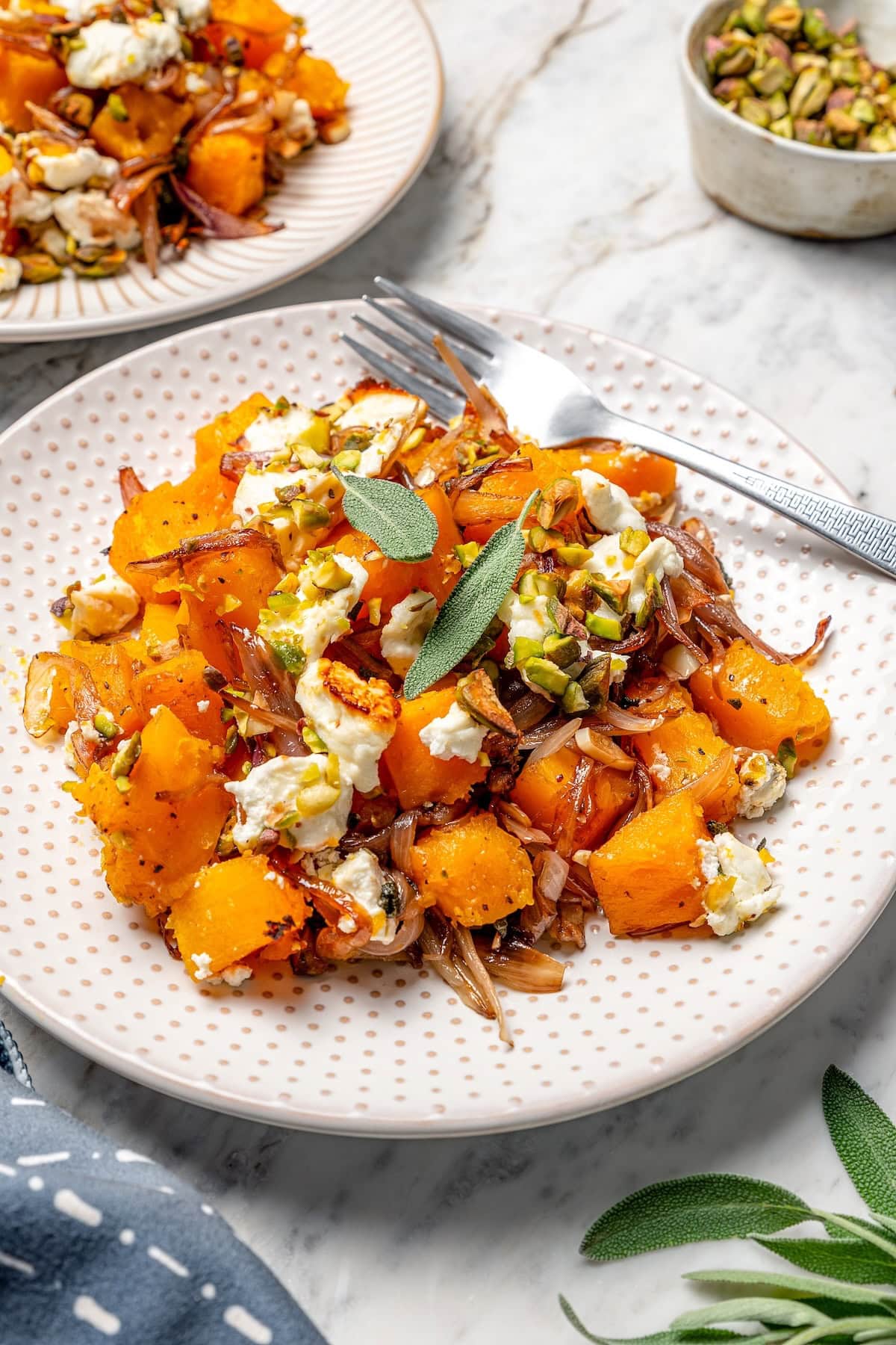Baked butternut squash is layered with creamy goat cheese, shallots, and pistachios in this cozy side dish recipe. Perfect for the holidays! Butternut squash bake on plate with goat cheese, pistachios, and sage.